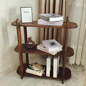 May include: A three-tiered, dark wooden bookshelf with rounded shelves. The top shelf holds a framed picture with the text "ENGLISH HOME" and two books. The middle shelf displays a decorative vase and more books. The bottom shelf has books and decorative items.