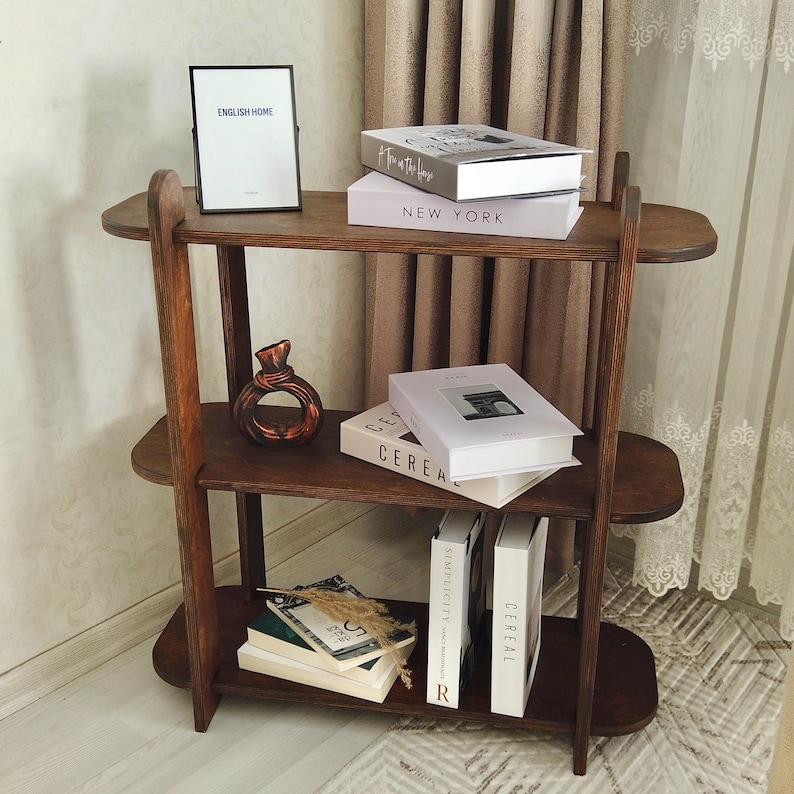 May include: A three-tiered, dark wooden bookshelf with rounded shelves. The top shelf holds a framed picture with the text "ENGLISH HOME" and two books. The middle shelf displays a decorative vase and more books. The bottom shelf has books and decorative items.