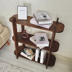 May include: A dark brown wooden bookshelf with three shelves. The top shelf holds a framed print with the text "ENGLISH HOME" and several books. The middle shelf has a decorative vase and more books. The bottom shelf has books and decorative items.