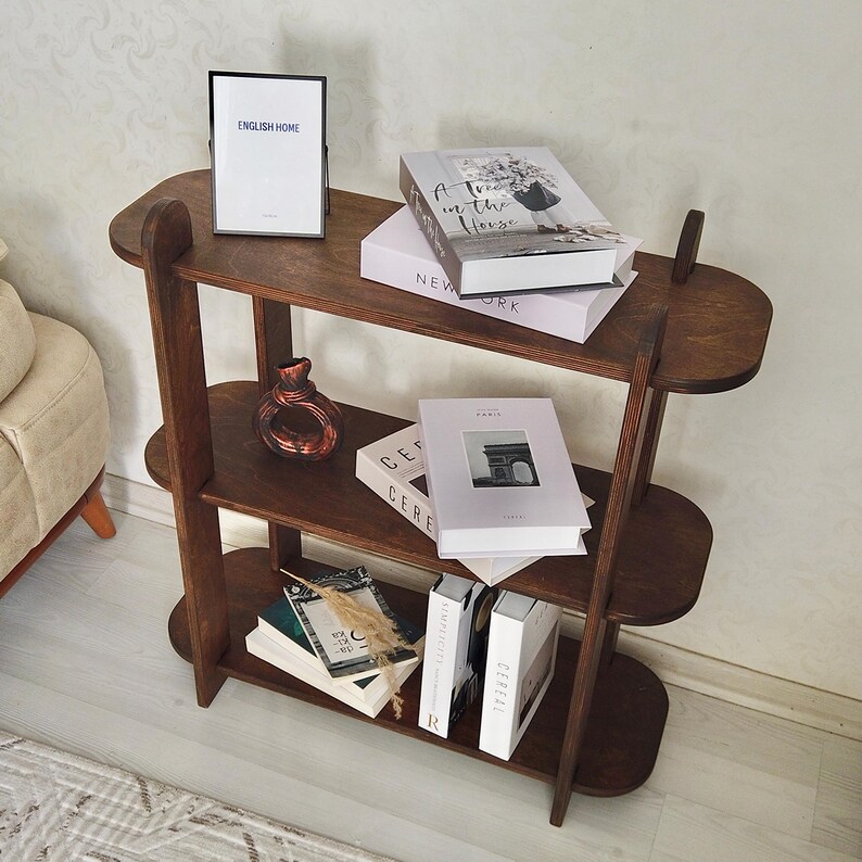 May include: A dark brown wooden bookshelf with three shelves. The top shelf holds a framed print with the text "ENGLISH HOME" and several books. The middle shelf has a decorative vase and more books. The bottom shelf has books and decorative items.