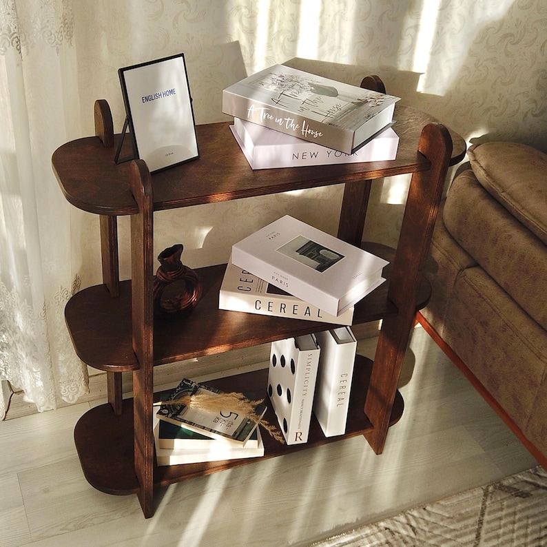 May include: A dark brown wooden bookshelf with three oval shelves. The top shelf holds books with the text "Tre in the House" and "NEW YORK". The middle shelf has books with the text "CEREAL" and "PARIS". A framed picture with the text "ENGLISH HOME" is on the top shelf.