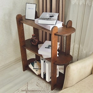 May include: A dark brown wooden bookshelf with three shelves and a unique curved design. The shelves hold books, decorative items, and a framed print that reads "ENGLISH HOME". The bookshelf is placed near a beige armchair.