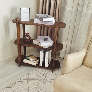 May include: A three-tiered, dark brown wooden bookshelf with oval shelves. The shelves hold books, a framed sign that reads "ENGLISH HOME", and a decorative vase. The bookshelf is placed near a patterned rug and a beige armchair.