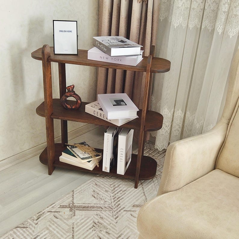 May include: A three-tiered, dark brown wooden bookshelf with oval shelves. The shelves hold books, a framed sign that reads "ENGLISH HOME", and a decorative vase. The bookshelf is placed near a patterned rug and a beige armchair.