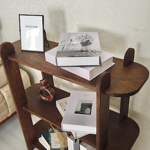 May include: A dark brown wooden bookshelf with three shelves. The top shelf holds a framed print with the text "ENGLISH HOME" and a stack of books. A decorative vase and more books are on the lower shelves.