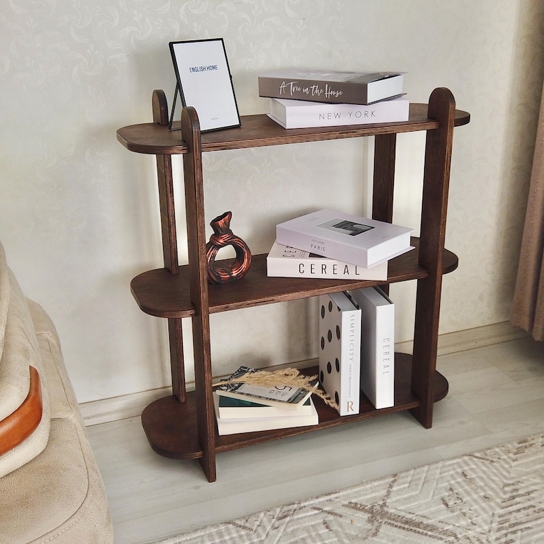 May include: A three-tiered, dark wood bookshelf with oval shelves. The top shelf holds a framed print with the text "ENGLISH HOME" and two books. The middle shelf displays a decorative vase and books. The bottom shelf has books and decorative items.
