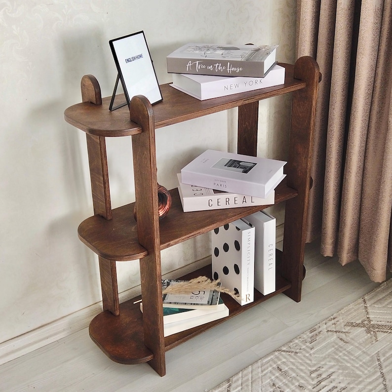 May include: A dark brown wooden bookshelf with three shelves. The top shelf holds a framed picture and two books. The middle shelf has two books, and the bottom shelf has books and decorative items. The bookshelf is against a light-colored wall.