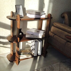May include: A three-tiered, dark wood bookshelf with rounded edges. The shelves hold books with titles like "NEW YORK" and "CEREAL", a framed picture, and a decorative sculpture. The bookshelf is in a room with a sofa.