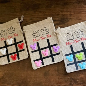 May include: Three burlap bags with Mickey Mouse hands printed on the front. Each bag has a tic-tac-toe board with black lines and colorful buttons in the squares. The text "Mic-Tac-Toe" is printed on each bag.