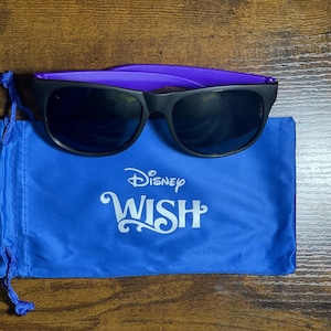 Puede incluir: Gafas de sol negras con montura morada y lentes oscuras sobre una bolsa azul con cordón. La bolsa presenta el logotipo de Disney WISH en blanco. Las gafas están sobre una superficie de madera.