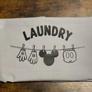 Puede incluir: Una bolsa de lavandería gris claro con la palabra "LAUNDRY" en letras mayúsculas negras grandes. Debajo, un tendedero muestra imágenes de dibujos animados de guantes, la cabeza de Mickey Mouse y pantalones cortos. La bolsa está doblada sobre una superficie de madera.