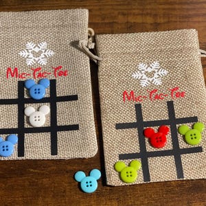 May include: Two burlap bags with a snowflake design and the text "Mic-Tac-Toe" printed on them. Each bag has a tic-tac-toe board with buttons in the shape of Mickey Mouse heads in blue, green, red, and white.
