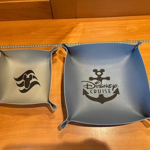 May include: Two gray and blue fabric trays with a Disney Cruise logo and a Disney Cruise line logo. The Disney Cruise logo is a black anchor with the words "Disney Cruise" underneath. The Disney Cruise line logo is a black Mickey Mouse head with three waves underneath.