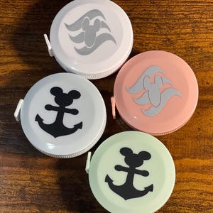 May include: Four round measuring tapes in pastel colors: white, pink, light green, and light gray. Each tape has a different design: a Mickey Mouse head with waves or a Mickey Mouse head with an anchor. The tapes are on a dark wood surface.