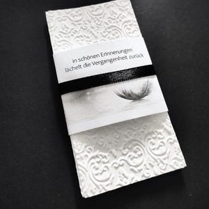 Könnte beinhalten: Eine weiße Karte mit geprägtem floralem Muster, umwickelt mit einem schwarzen Band. Die Karte enthält den deutschen Text: "in schönen Erinnerungen lächelt die Vergangenheit zurück" und ein Graustufenbild einer Feder.
