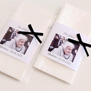 Könnte beinhalten: Zwei weiße Papierservietten mit einer weißen Papierbanderole, bedruckt mit einem Schwarz-Weiß-Foto einer älteren Person, dem Text "Du fehlst", dem Namen "Martina" und einem Datum, verziert mit einer kleinen schwarzen Schleife.