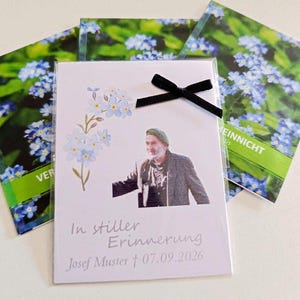 Könnte beinhalten: Eine rechteckige Gedenkkarte mit einem Foto einer Person mit grünem Barett und dunkler Jacke, verziert mit hellblauen Vergissmeinnicht-Blüten, einer kleinen schwarzen Schleife und dem Text In stiller Erinnerung Josef Muster 07.09.2026.
