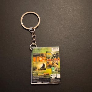 Könnte beinhalten: Ein rechteckiger Schlüsselanhänger mit der Rückseite einer Videospielhülle. Die Hülle hat ein farbenfrohes Design mit Bildern von Charakteren und Szenen aus dem Spiel. Der Schlüsselanhänger enthält einen silbernen Metallring und eine Kette.