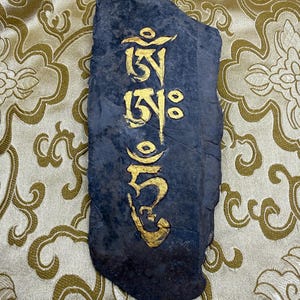May include: A dark gray stone with gold-colored Tibetan script. The stone has a rough, natural texture and is rectangular with rounded edges. The gold script is vertically aligned and appears to be hand-painted.