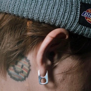 Può includere: Una persona con un berretto grigio con un logo sul retro ha un tatuaggio di smiley pixelato dietro l'orecchio. Indossa un orecchino a cerchio d'argento.