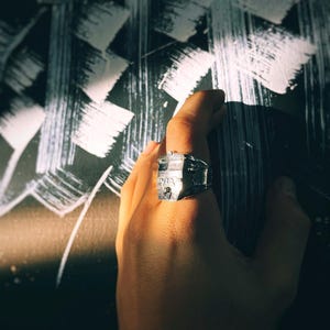 May include: A silver ring with a detailed design of a building. The ring is worn on a person's finger.