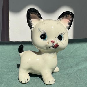 Può includere: Statuetta vintage a forma di gatto in ceramica con corpo color crema, orecchie e coda nere e occhi blu. Il gatto ha la lingua rossa di fuori ed è in posa giocosa. Probabilmente un oggetto decorativo.