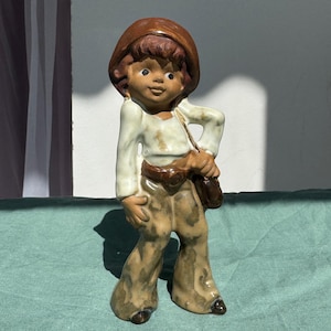 Può includere: Statuina in ceramica di un giovane con cappello marrone, camicia bianca e pantaloni a zampa. La statuina ha una borsa a tracolla e una cintura marrone. La figura è su una superficie verde.