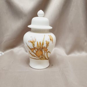 May include: A white ceramic jar with a lid, featuring a floral design in shades of orange and brown. The jar has a rounded body and a lid with a small knob. The floral design includes irises and a butterfly.