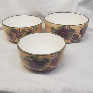 May include: Three ceramic bowls with a beige exterior, white interior, and a green rim. Each bowl is decorated with a purple grape cluster design. The bowls are of varying sizes and are arranged together.