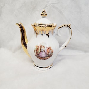 May include: White porcelain teapot with gold trim and a lid. The teapot features a handle, spout, and a decorative scene of people in a garden. The gold trim is applied to the spout, handle, lid, and the top of the teapot.