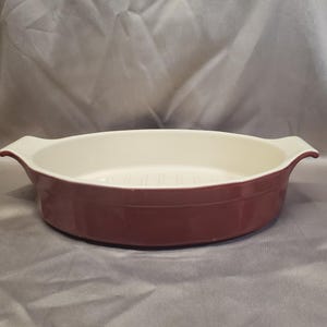 May include: Oval-shaped burgundy baking dish with a white interior. The dish has two small handles on either side. The interior is white with a ridged pattern. This dish is suitable for baking and serving.
