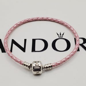 May include: A light pink braided leather bracelet featuring a central silver-toned metal charm engraved with the letters P, A, N, D, O, R, A. The bracelet includes two silver-toned metal end caps that secure the braided cord to the central charm.