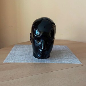 Op de afbeelding: Een glanzende zwarte keramische hoofdsulptuur met duidelijke gelaatstrekken, waaronder een neus, mond en oren. De sculptuur staat op een grijze placemat, tegen een lichtbruine achtergrond. Het hoofd is een decoratief item.