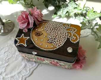 Op de afbeelding: Kleine, rechthoekige houten doos met een donkerbruine afwerking. De deksel heeft een maanontwerp met een gezicht, versierd met strass steentjes. Een ster en stof met bloemenpatroon sieren de doos. Roze bloemen en heldere strass steentjes zijn rond de doos geplaatst.