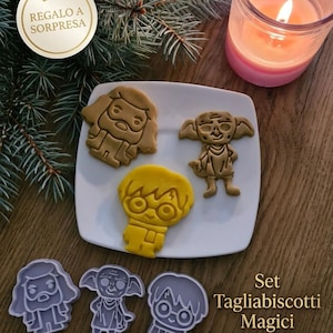 Puede incluir: Un plato blanco exhibe tres galletas con forma de personajes, incluyendo un mago, un elfo doméstico y un chico con gafas. Debajo, se ven tres cortadores de galletas con las mismas formas. El texto "Set Tagliabiscotti Magici" y "3 Formine incluse" también son visibles.