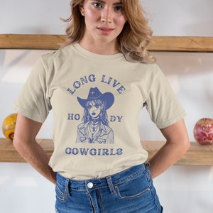Puede incluir: Una camiseta beige con un gr&aacute;fico azul de una mujer con un sombrero de vaquero y el texto "Long Live Holy Cowgirls".