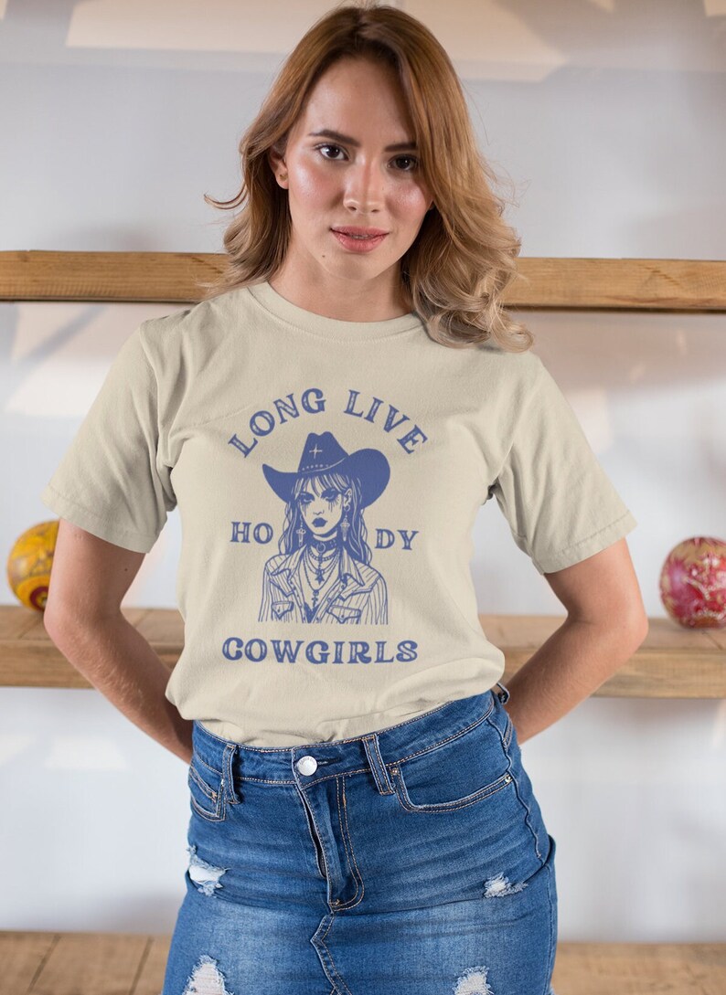 Puede incluir: Una camiseta beige con un gr&aacute;fico azul de una mujer con un sombrero de vaquero y el texto "Long Live Holy Cowgirls".