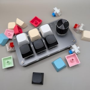 May include: A clear plastic panel with a knob and eight colourful keycaps. The keycaps are arranged in a grid pattern and are black, white, cream, pink, blue, and light green. The panel is on a grey surface.
