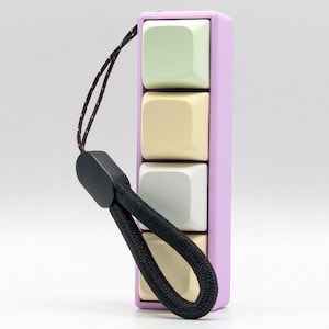 Op de afbeelding: Een lavendelkleurige sleutelhanger met een zwart koord en vier kleurrijke vierkante knoppen. De knoppen zijn lichtgroen, geel, grijs en beige.