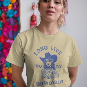 Puede incluir: Una camiseta beige claro con un gr&aacute;fico azul de una vaquera con un sombrero y una cruz. El texto "Long Live Cowgirls" est&aacute; impreso en azul en la camiseta.