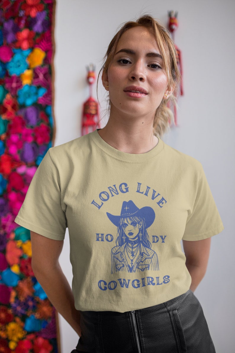 Puede incluir: Una camiseta beige claro con un gr&aacute;fico azul de una vaquera con un sombrero y una cruz. El texto "Long Live Cowgirls" est&aacute; impreso en azul en la camiseta.