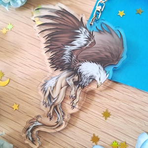 May include: A clear acrylic keyring depicting a griffin with brown and white feathers, a tan body, and a curved tail. The keyring has a gold-coloured chain and is decorated with gold star-shaped confetti.