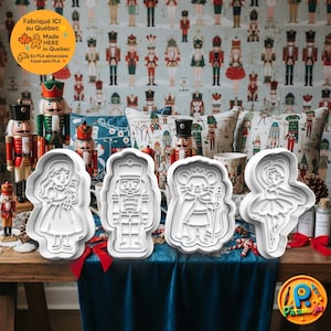 May include: Four white plastic cookie cutters shaped like a girl, a nutcracker, a mouse, and a ballerina are on a wooden table. The background shows nutcracker figures, pillows, and wallpaper. The image includes text that says "Made HERE in Quebec."