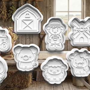 May include: A selection of white plastic cookie cutters in farm animal and barnyard shapes, including a tractor, barn, farmer, windmill, chick, pig, sheep, and cow. The background features a wooden table and a sign that says "La Ferme du Bonheur."