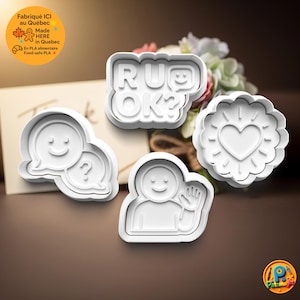 May include: Four white cookie cutters with various designs. One cutter spells "RU OK?" with a smiley face. Other cutters include a heart with sun rays, a person waving, and a speech bubble with a question mark and a smiley face.