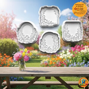 May include: Four white floral-themed cookie cutters in various shapes: square, rectangular, and oval. The cutters are arranged against a blurred outdoor background with a wooden picnic table and a vase of flowers. The text "Fabriqué ICI au Québec Made HERE in Quebec" is visible.