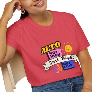 May include: A red t-shirt with a graphic design that says "Alto, Not High, Just Right, Not Low" with a sun and musical notes.