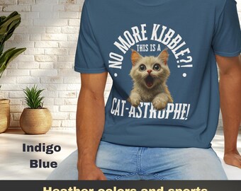 May include: A man wearing a blue t-shirt with a white graphic of a cat and the text "No More Kibble? This is a Cat-Astrophy!"  Heather colors and sports grey include polyester.  NO MORE KIBBLE CAT-ASTROPHE craftyharmonies.etsy.com