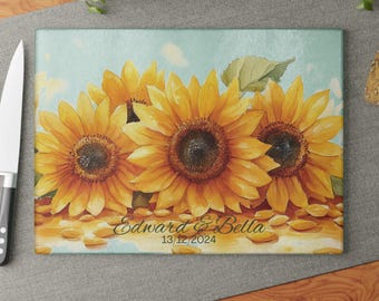 Peut inclure: Une planche à découper en verre personnalisée avec un fond bleu et trois tournesols jaunes. Le texte "Edward & Bella 13/12/2024" est imprimé sur la planche.