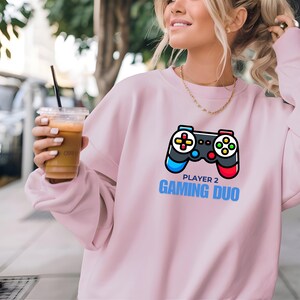May include: Pink sweatshirt with a graphic of a video game controller and the text "Player 2 Gaming Duo".